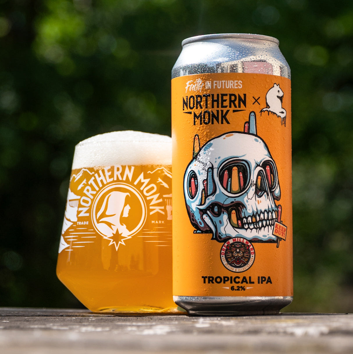 A can and a filled glass of Northern Monk Tropical IPA beer, with the branded orange label clearly visible, set on a wooden surface against a blurred green outdoor background.