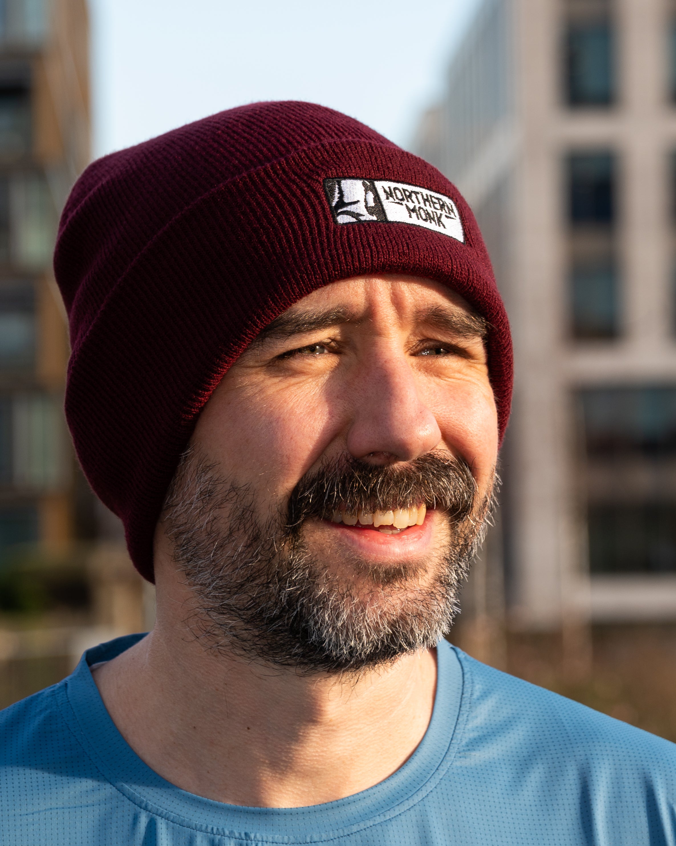 Man wearing a burgundy beanie with a logo, smiling outdoors.
